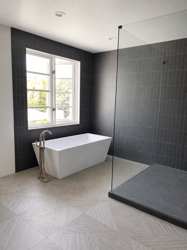 Graphite vertical stack-bond porcelain shower with a floating corner bench, recessed niche, brushed brass rainhead and a wood-look chevron tile floor