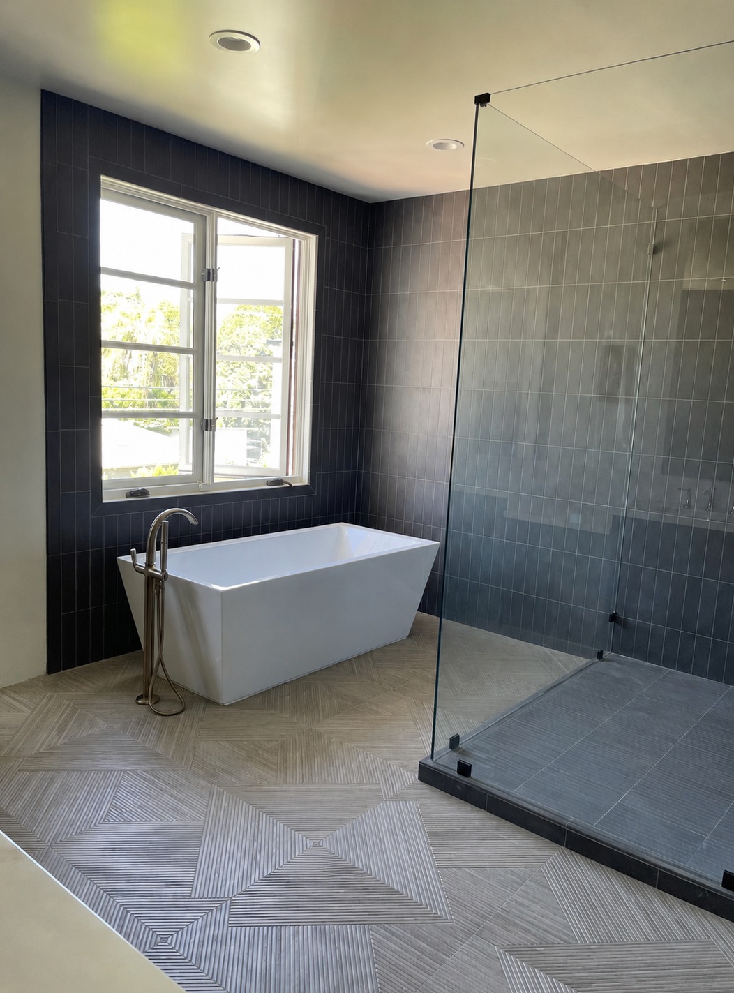 Primary bathroom with charcoal stack-bond vertical wall tile, a patterned porcelain floor, a freestanding soaking tub and a frameless glass curbless shower, installed by Horizon Tile Professional